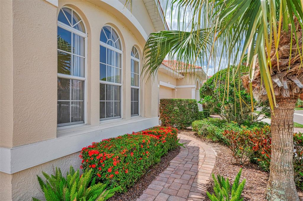 A paved walkway leads past a tan house with arched windows and red flowering hedges — 7803 33RD STREET E, SARASOTA, FL