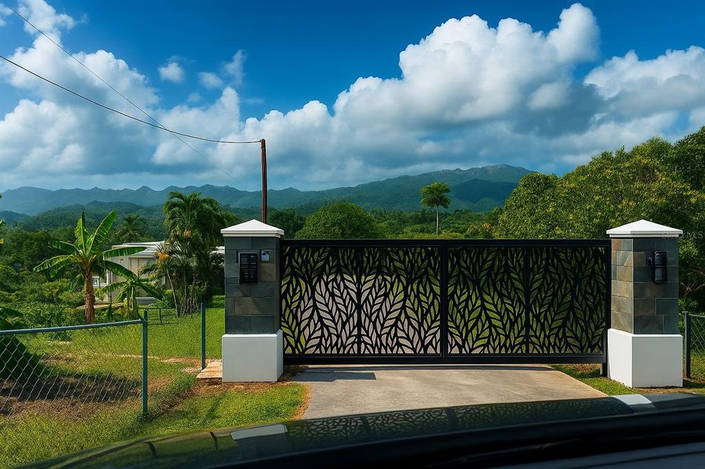 A decorative black metal gate stands between two stone pillars with a mountain view background — SOLAR 39 BO FRONTON, CIALES, PR