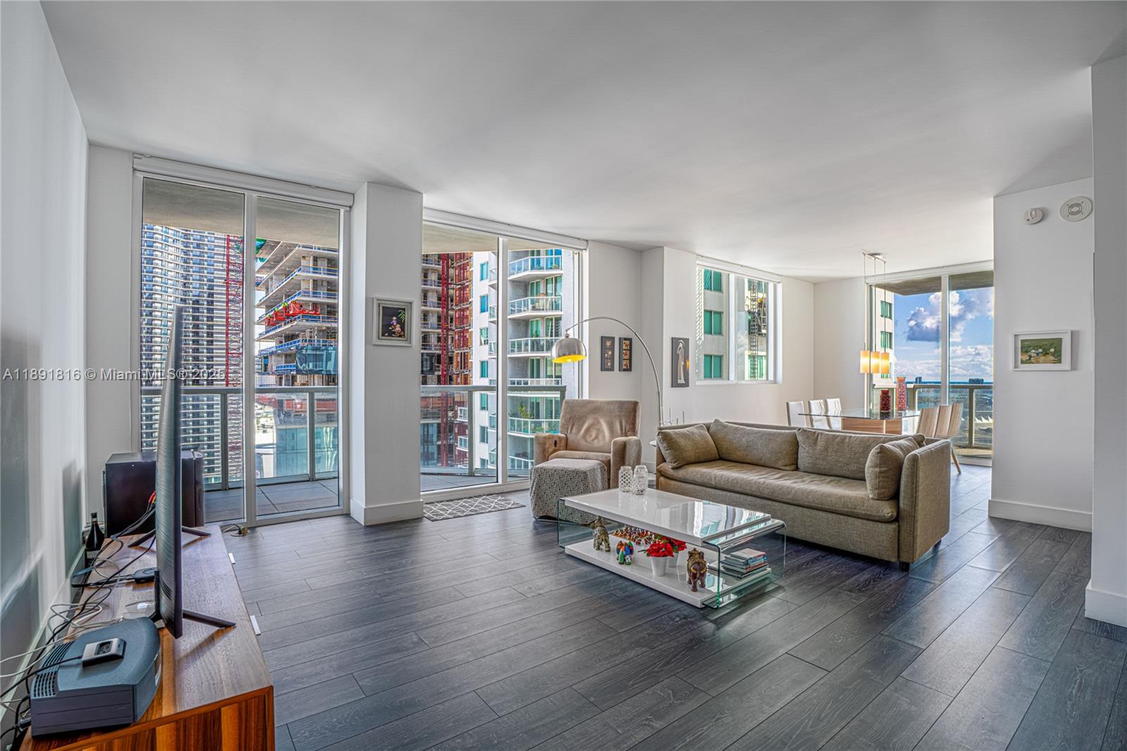 A living room with dark wood floors, a beige sofa, and floor-to-ceiling windows overlooking construction — 253 NE 2nd St # 3203, Miami FL 33132, MIAMI, FL
