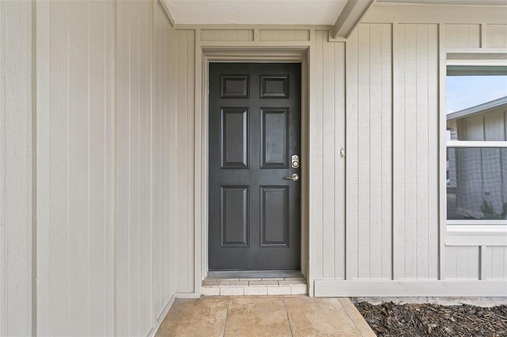 A dark gray six-panel front door is set within a light-colored vertical siding exterior — 8022 IDLEBRIAR LANE, PORT RICHEY, FL