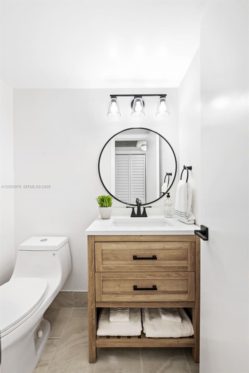 A modern bathroom features a wood vanity, round mirror, and white toilet on tiled flooring — 17 Lyndhurst # A, Deerfield Beach FL 33442, DEERFIELD BEACH, FL