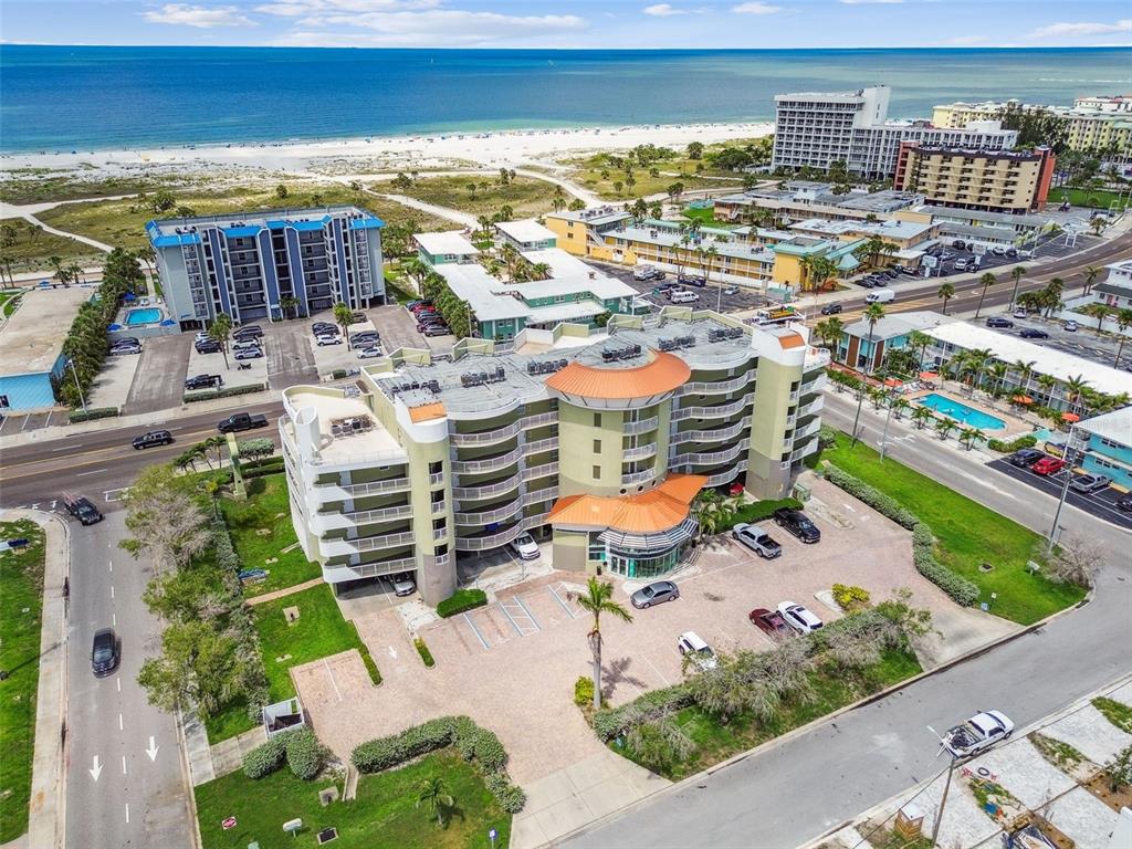 An aerial view shows a multi-story apartment building with a parking lot near the beach — 11605 GULF BOULEVARD UNIT 506, TREASURE ISLAND, FL