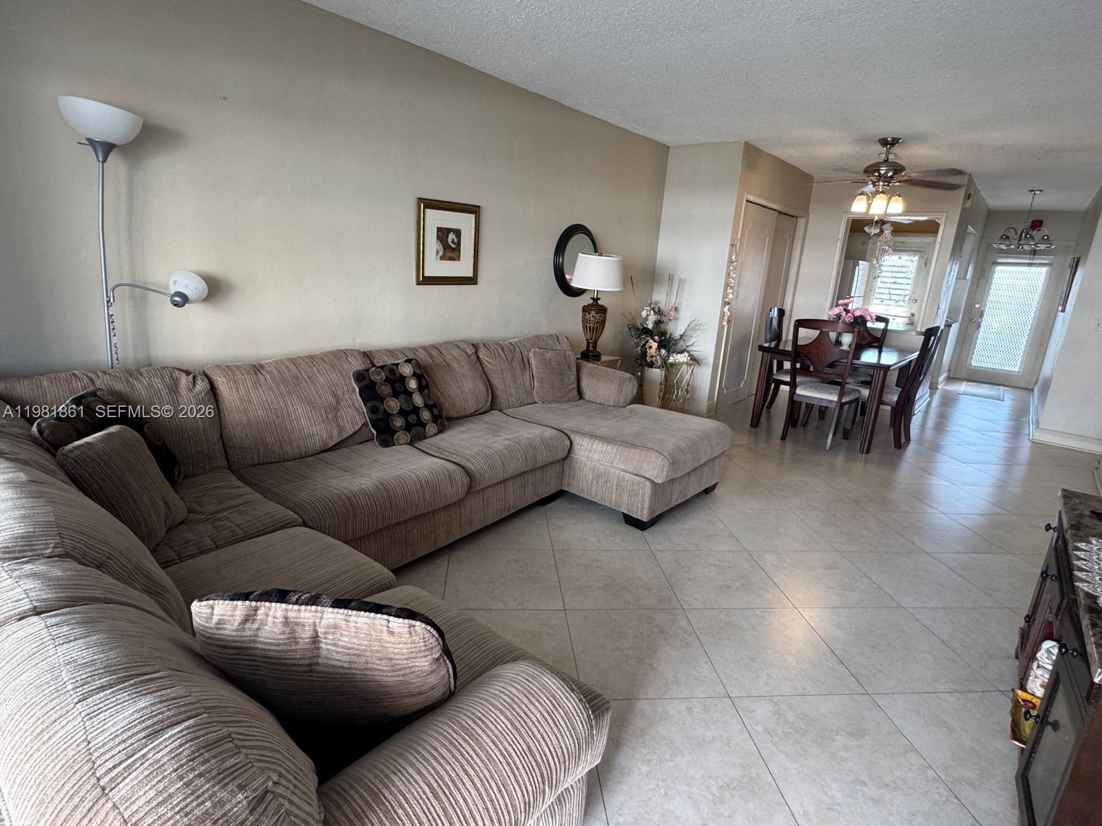 A living room with a large sectional sofa and tile flooring leads to a dining area — 450 Paradise Isle Blvd # 209, Hallandale Beach FL 33009, HALLANDALE BEACH, FL