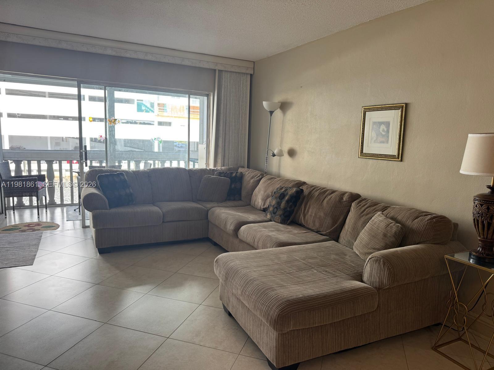 A living room features a large beige sectional sofa, tile flooring, and a sliding glass door — 450 Paradise Isle Blvd # 209, Hallandale Beach FL 33009, HALLANDALE BEACH, FL