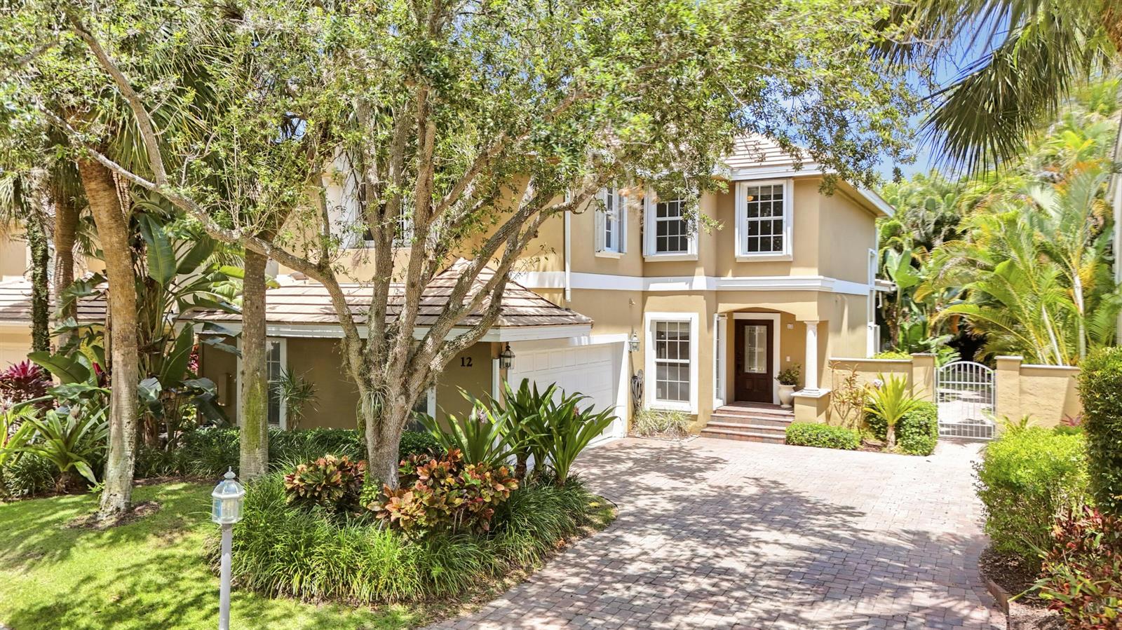 A two-story tan house with a brick driveway, white garage door, and lush tropical landscaping — 12 Grand Bay Circle, Juno Beach FL 33408, JUNO BEACH, FL