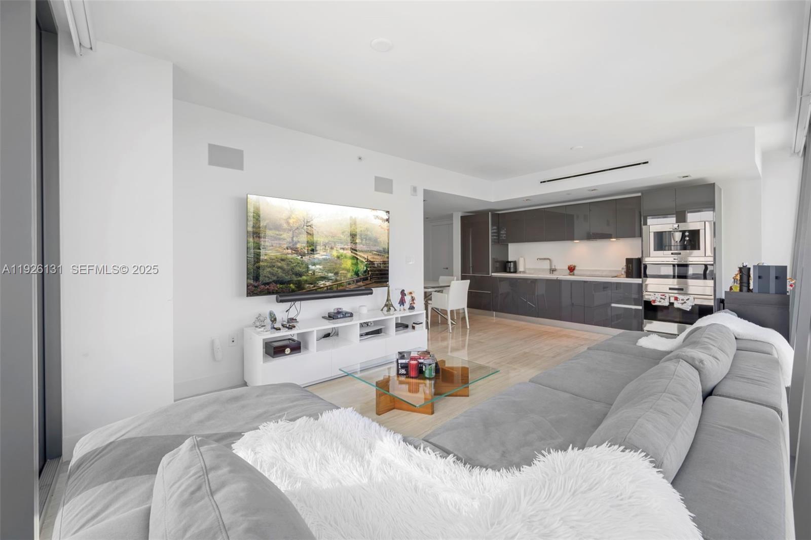 A modern living room features a gray sectional sofa, a wall-mounted television, and a kitchen — 1451 Brickell Ave # 1106, Miami FL 33131, MIAMI, FL