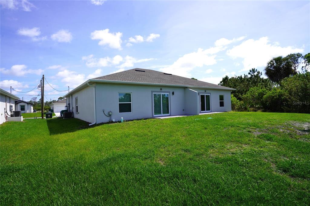 The rear exterior of a light-colored single-story house features a grassy yard and sliding doors — 12030 TETZEL AVENUE, PORT CHARLOTTE, FL