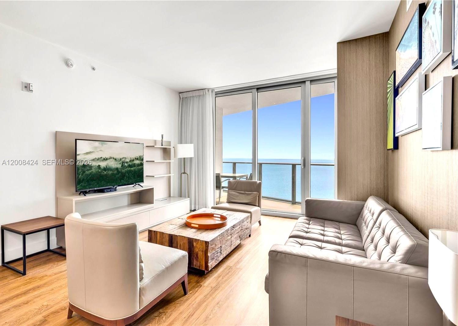A modern living room features a leather sofa, wood coffee table, and floor-to-ceiling ocean views — 4111 S Ocean Dr # 2303, Hollywood FL 33019, HOLLYWOOD, FL
