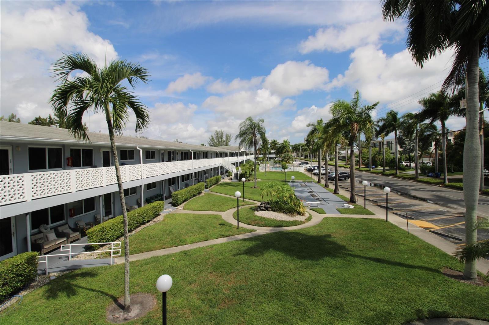 An exterior view of a multi-story apartment complex with a grassy courtyard and palm trees — 600 Layne Boulevard # 119, Hallandale Beach FL 33009, HALLANDALE BEACH, FL