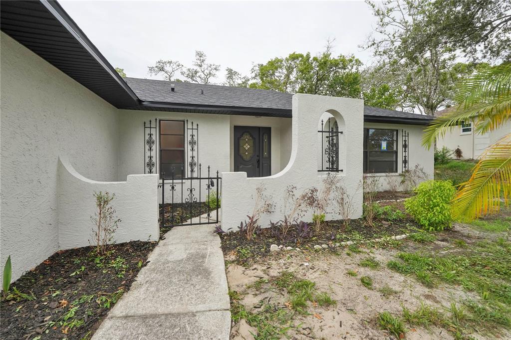 A single-story house exterior features a stucco facade, a gated courtyard, and a dark front door — 8081 WOODEN DRIVE, SPRING HILL, FL