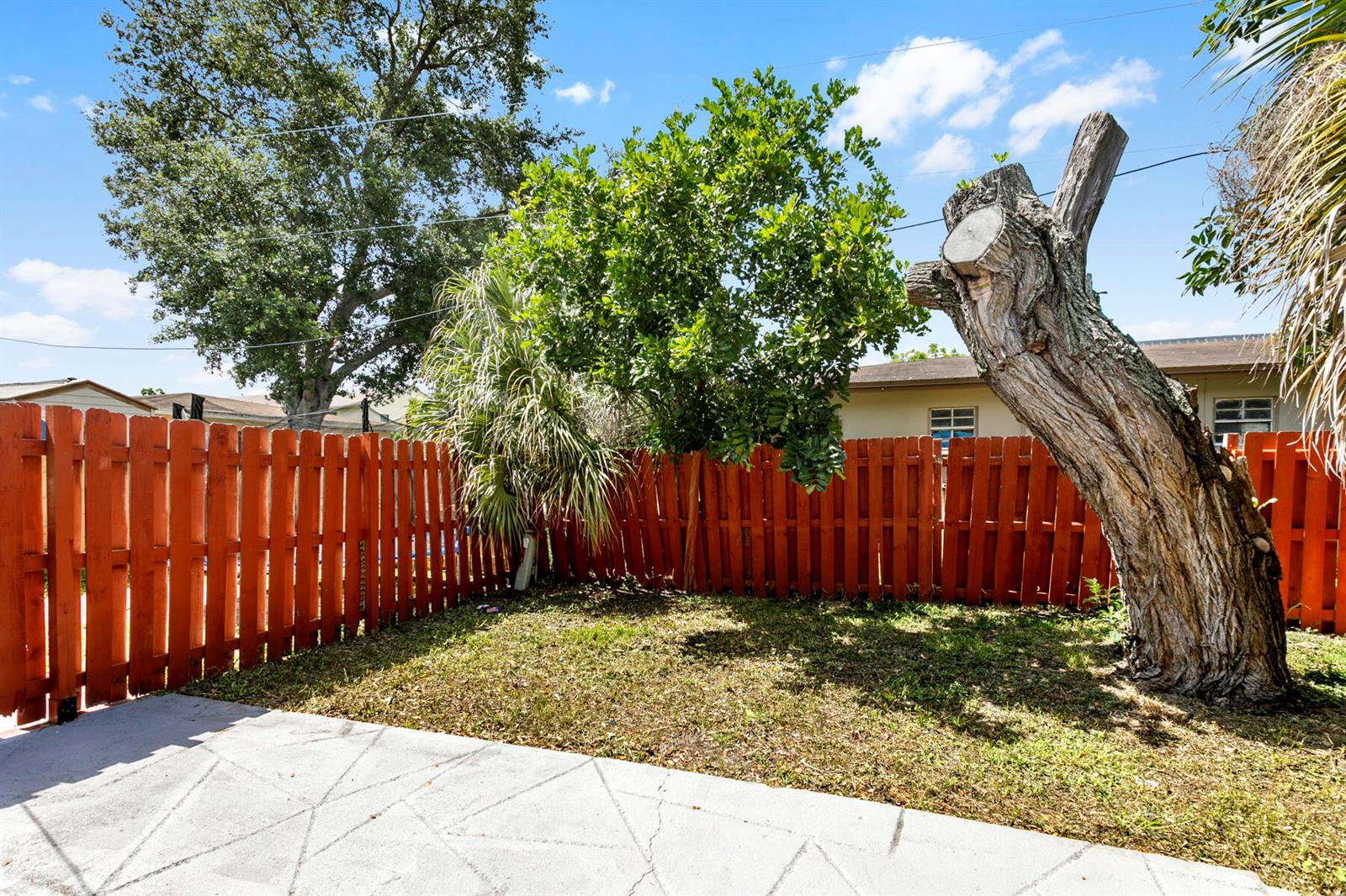 A backyard features a concrete patio, a red wooden fence, and a large tree trunk — RIVIERA BEACH, FL