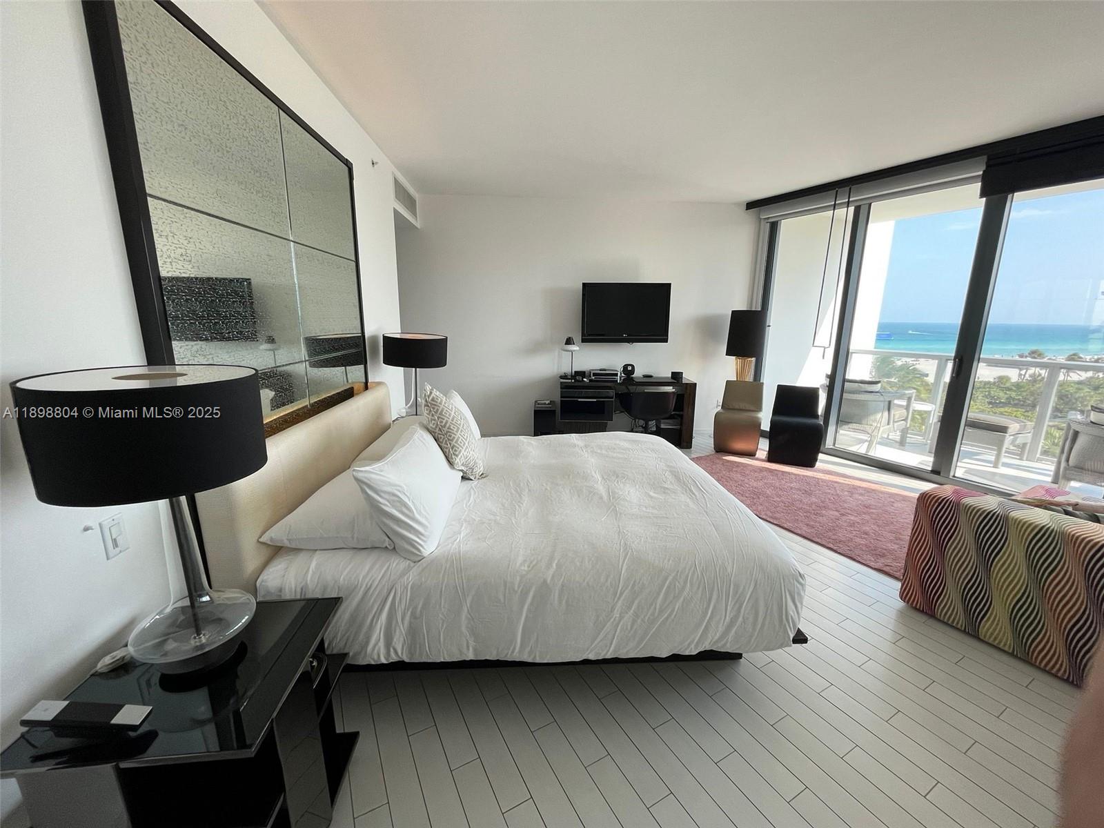 A modern bedroom features a king bed, mirrored wall, and floor-to-ceiling windows overlooking the ocean — 2201 Collins Ave # 626, Miami Beach FL 33139, MIAMI BEACH, FL