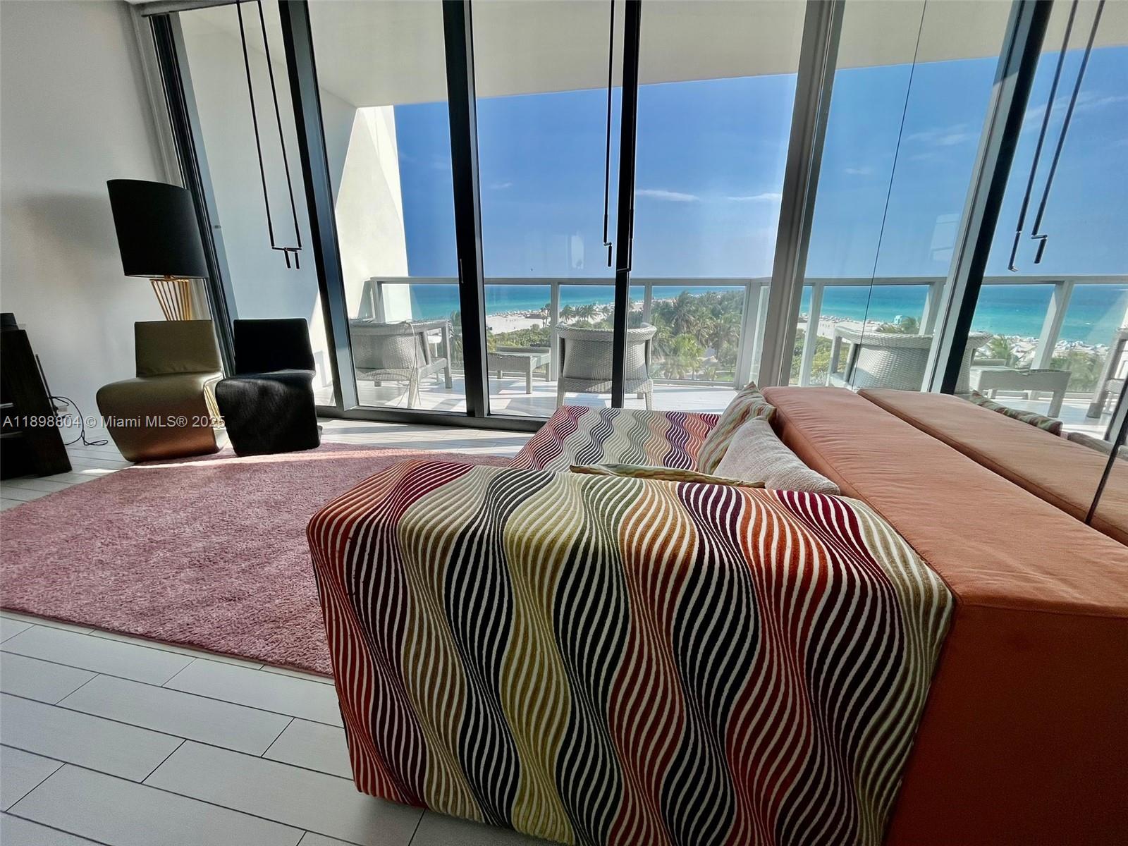 A modern room features a striped sofa, pink rug, and floor-to-ceiling windows overlooking the ocean — 2201 Collins Ave # 626, Miami Beach FL 33139, MIAMI BEACH, FL