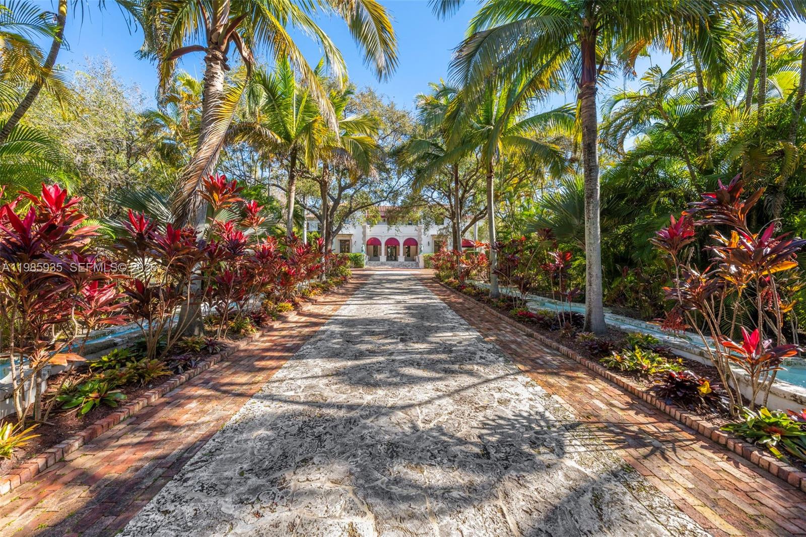 A long stone driveway lined with palm trees and red-leafed plants leads to a villa — 9401 Journeys End Rd, Coral Gables FL 33156, CORAL GABLES, FL