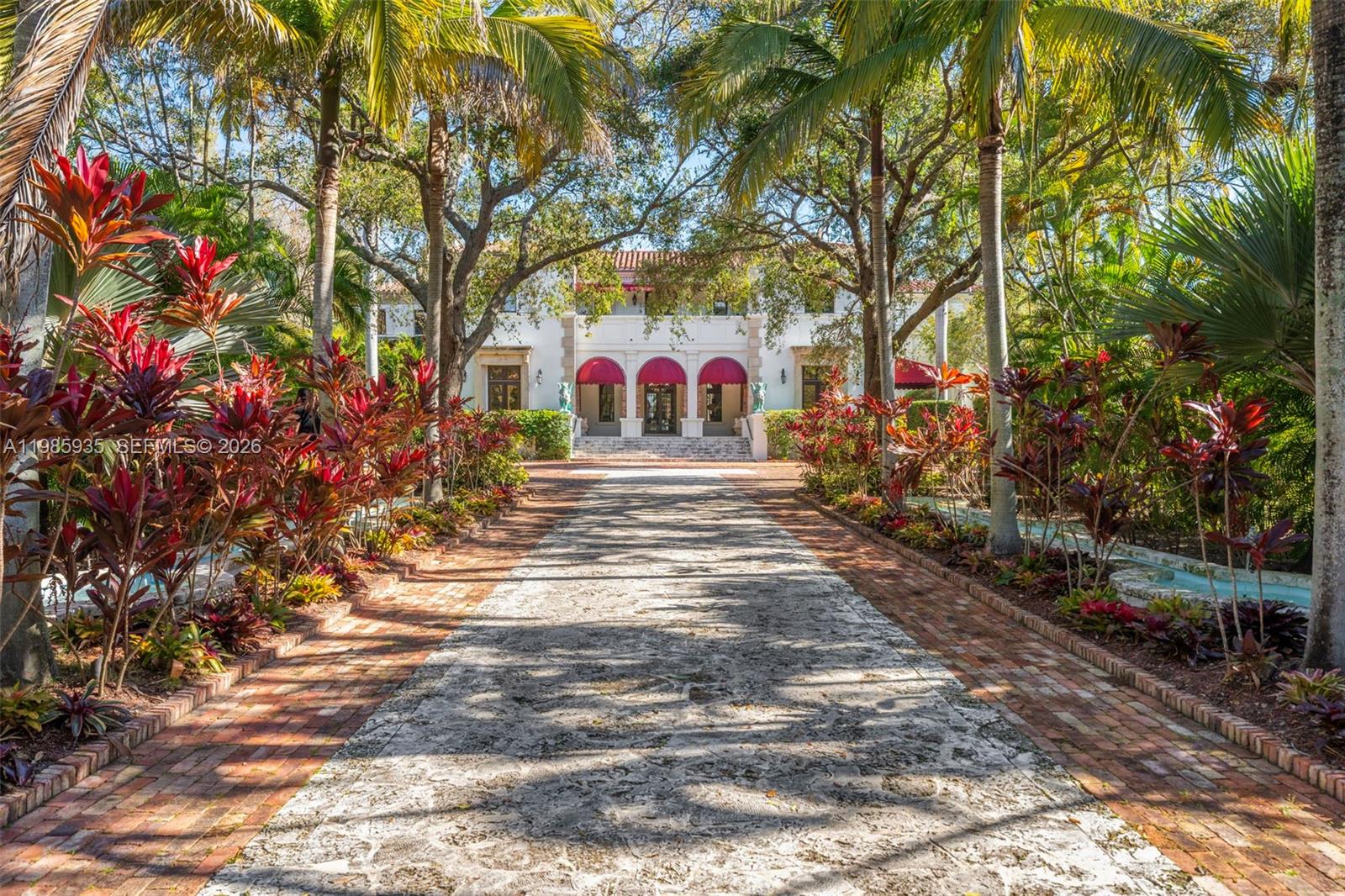 A long stone driveway leads to a white Mediterranean-style house with red awnings and landscaping — 9401 Journeys End Rd, Coral Gables FL 33156, CORAL GABLES, FL