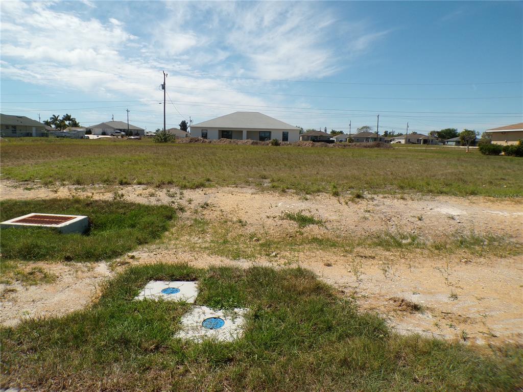 A vacant, grassy residential lot features a storm drain and utility access covers in the foreground — 1816 NE 8TH PLACE, CAPE CORAL, FL
