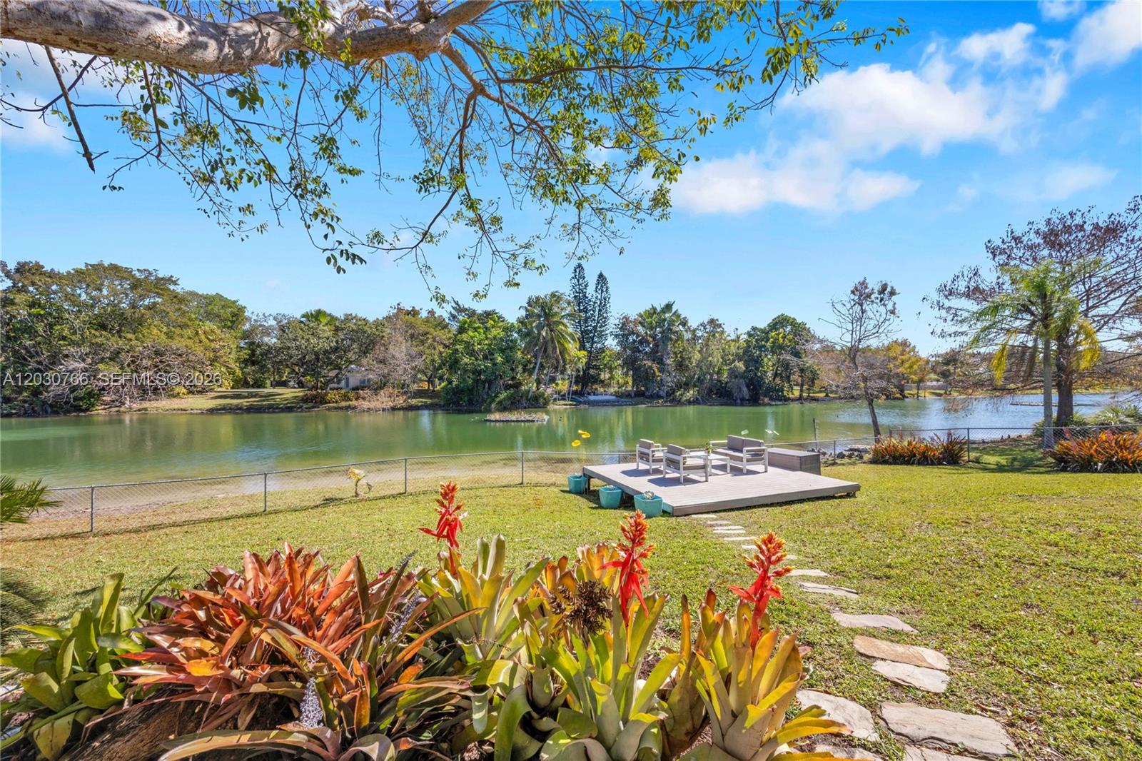 A backyard features a wooden deck with chairs overlooking a lake and lush landscaping — 11625 SW 70th Ave, Pinecrest FL 33156, PINECREST, FL