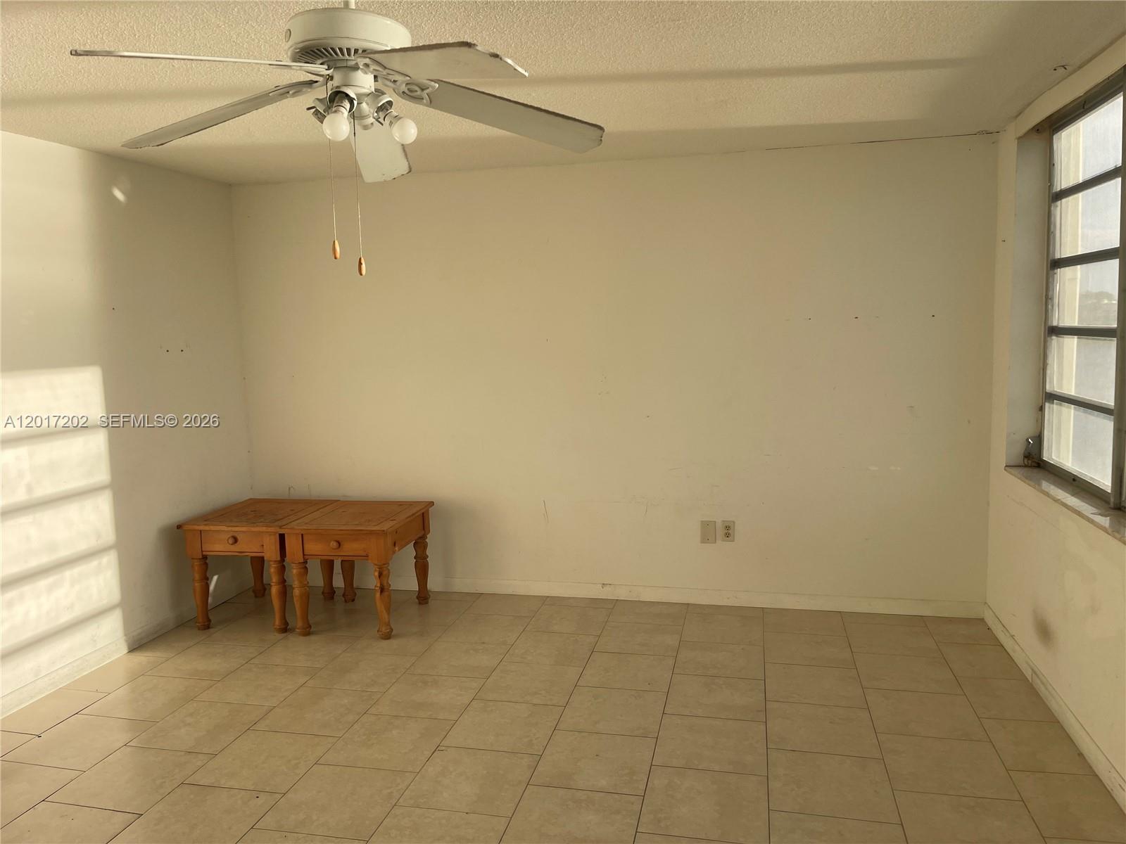 An empty room features white walls, beige tile flooring, a ceiling fan, and a wooden table — 1660 NE 191st St # 1, Miami FL 33179, MIAMI, FL