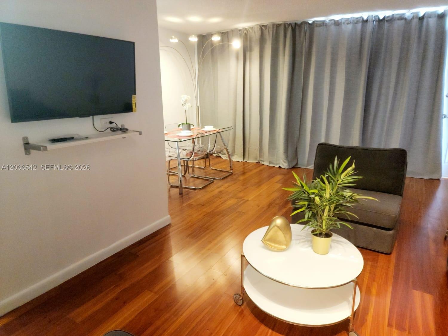 A living and dining area features hardwood floors, a wall-mounted television, and floor-to-ceiling curtains — 1100 Alton Rd # 3C, Miami Beach FL 33139, MIAMI BEACH, FL