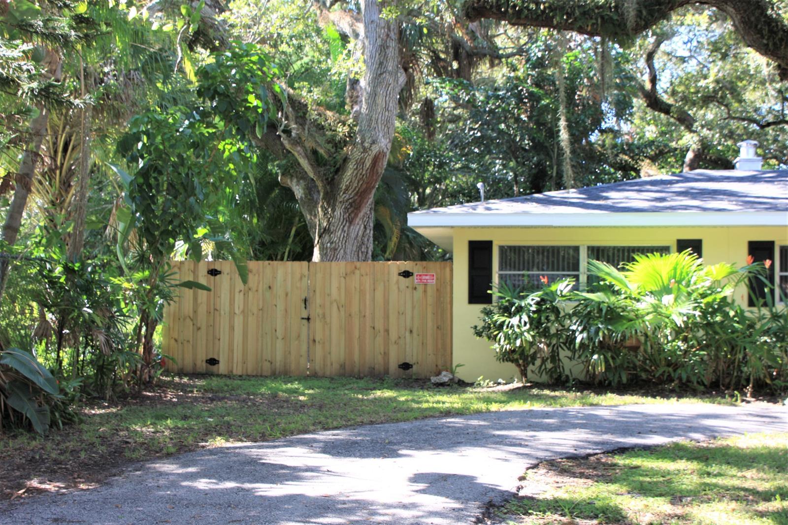 A wooden gate stands next to a yellow house with a driveway and lush trees — 635 18th Place, Vero Beach FL 32960, VERO BEACH, FL