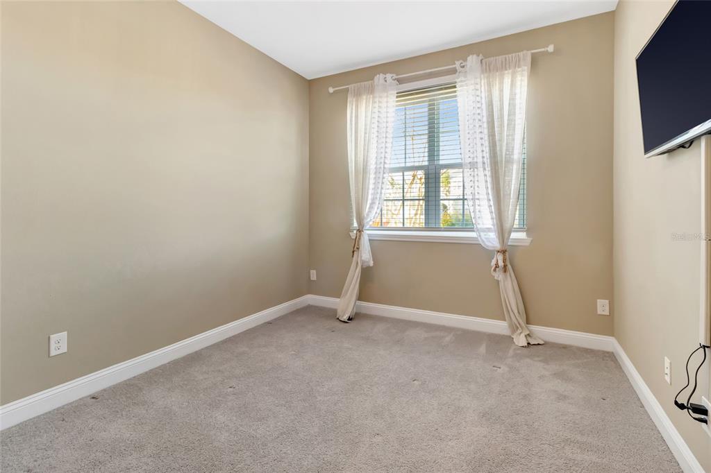 A bedroom features beige walls, gray carpeting, a window with white curtains, and a television — 549 CHAMPION RIDGE DRIVE, DAYTONA BEACH, FL