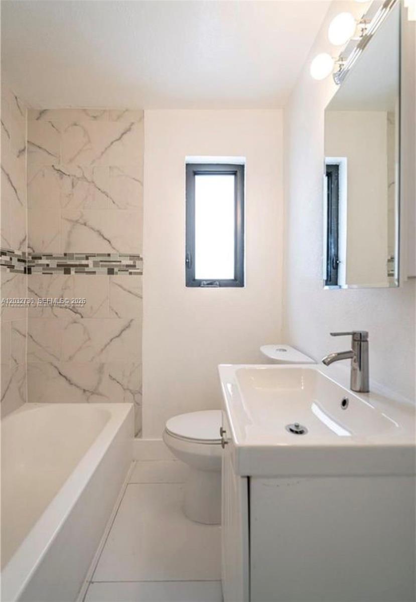 A bathroom features a white vanity, a toilet, and a bathtub with marble-patterned tile walls — 7935 Crespi Blvd # 3, Miami Beach FL 33141, MIAMI BEACH, FL