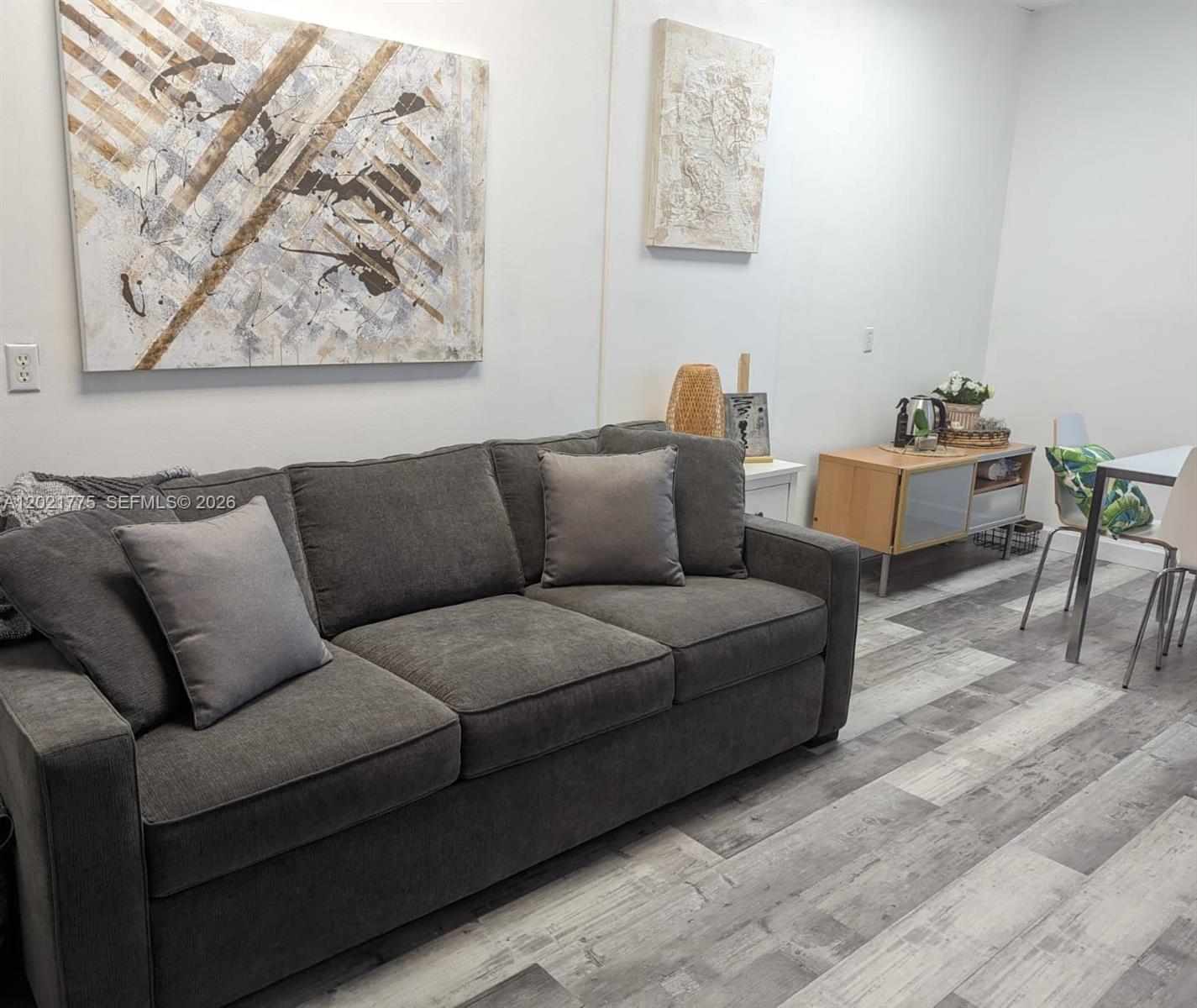 A living room features a gray sofa, abstract wall art, and light wood-look flooring — 12550 Biscayne Blvd # 220, North Miami FL 33181, NORTH MIAMI, FL
