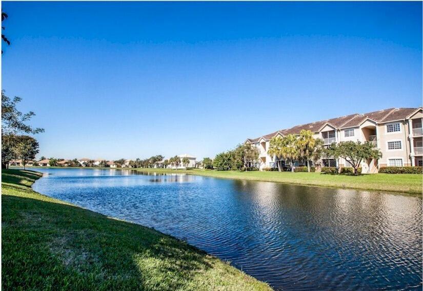 A multi-story apartment building sits beside a large lake under a clear blue sky — 8935 Okeechobee Boulevard # 204, West Palm Beach FL 33411, WEST PALM BEACH, FL