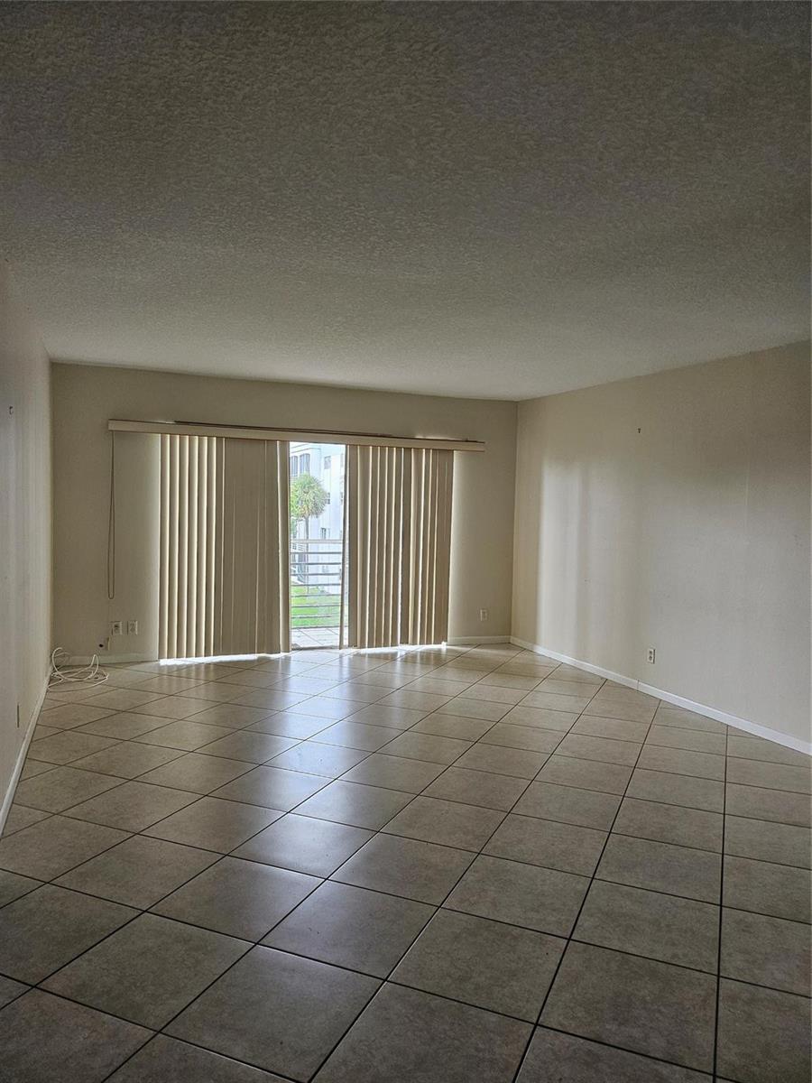An empty living room features beige tile flooring, light-colored walls, and vertical blinds over a balcony — 608 NE 2nd Street # 341, Dania Beach FL 33004, DANIA BEACH, FL