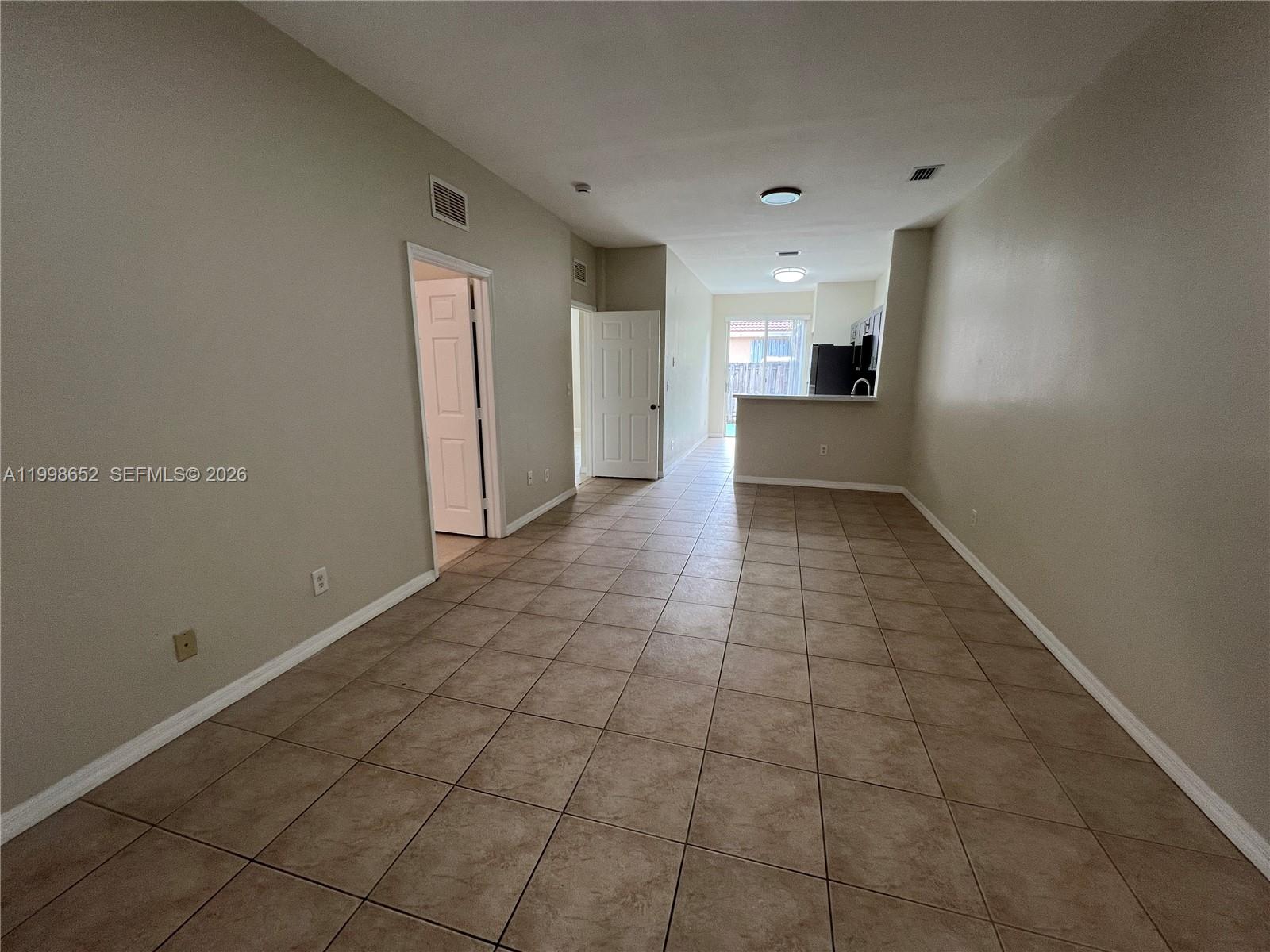 An empty living area with beige tile floors leads to a kitchen and sliding glass doors — 85 SW 16 AV, Homestead FL 33030, HOMESTEAD, FL