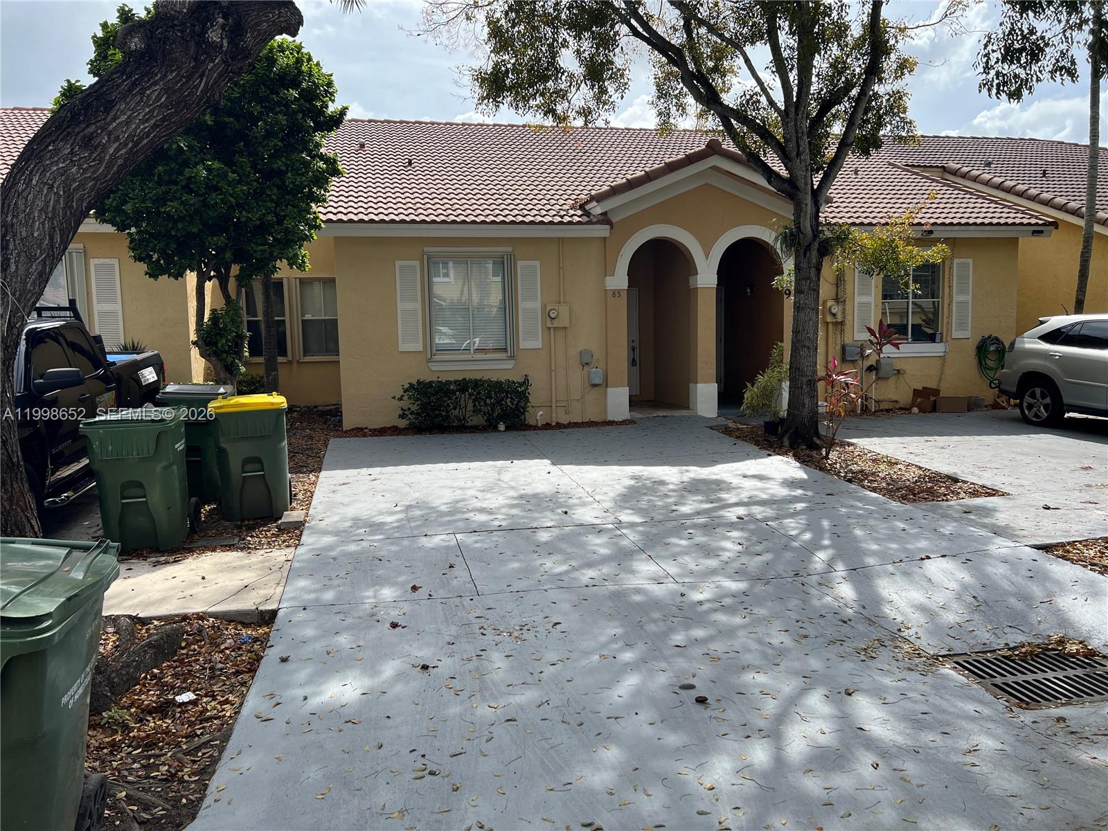 A single-story tan house features a gray concrete driveway, arched entryway, and two trash bins — 85 SW 16 AV, Homestead FL 33030, HOMESTEAD, FL