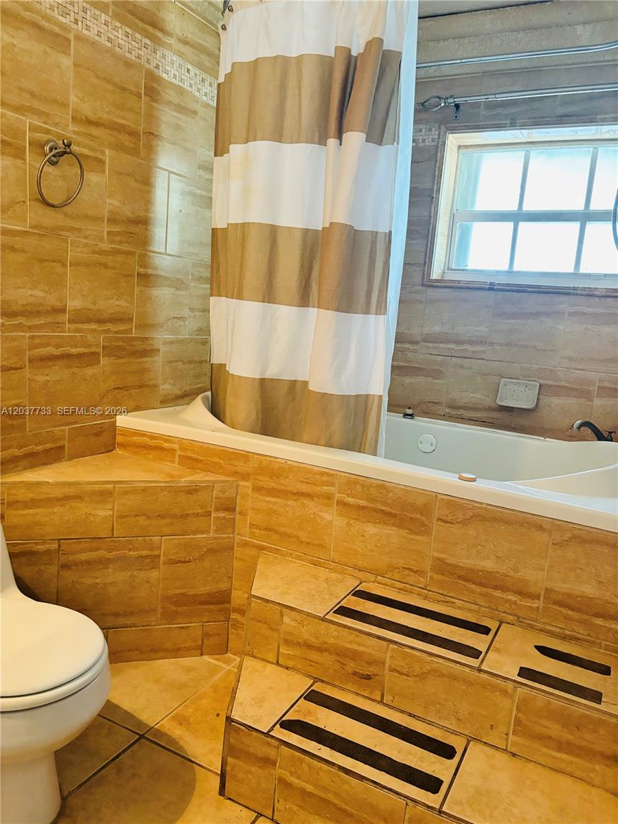 A bathroom features a tiled soaking tub with steps, a shower curtain, and a toilet — DAVIE, FL
