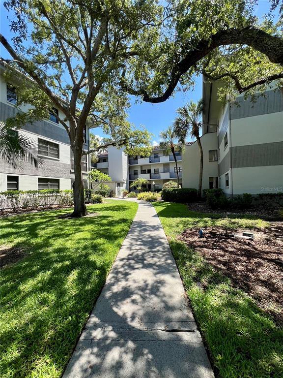 A concrete walkway leads between two multi-story apartment buildings surrounded by trees and grass — 2650 COUNTRYSIDE BOULEVARD UNIT B302, CLEARWATER, FL