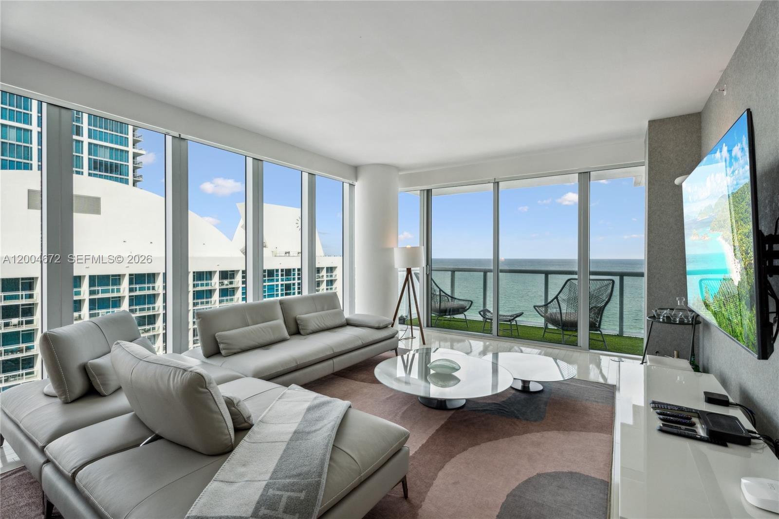 A modern living room features a grey sectional sofa, glass coffee table, and floor-to-ceiling windows — 6799 Collins Ave # 1602, Miami Beach FL 33141, MIAMI BEACH, FL