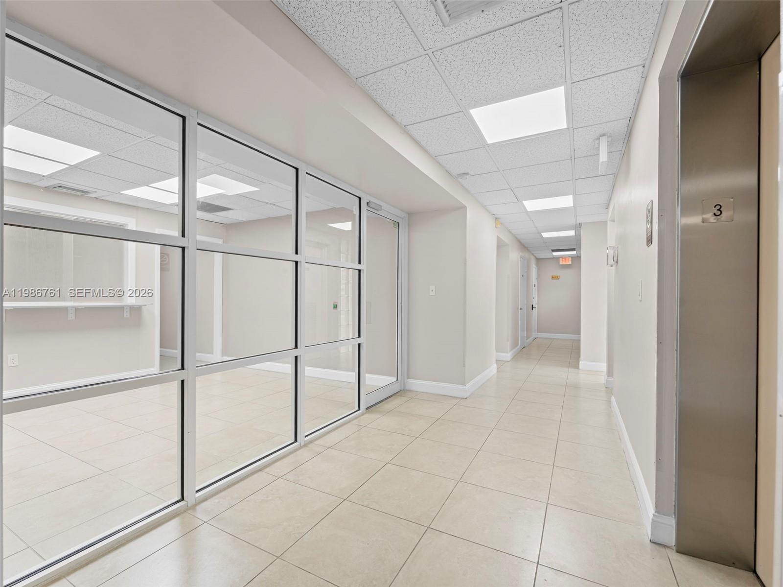 A hallway with tile flooring leads past a glass-walled office to an elevator — 1312 SW 27th Ave, Miami FL 33145, MIAMI, FL