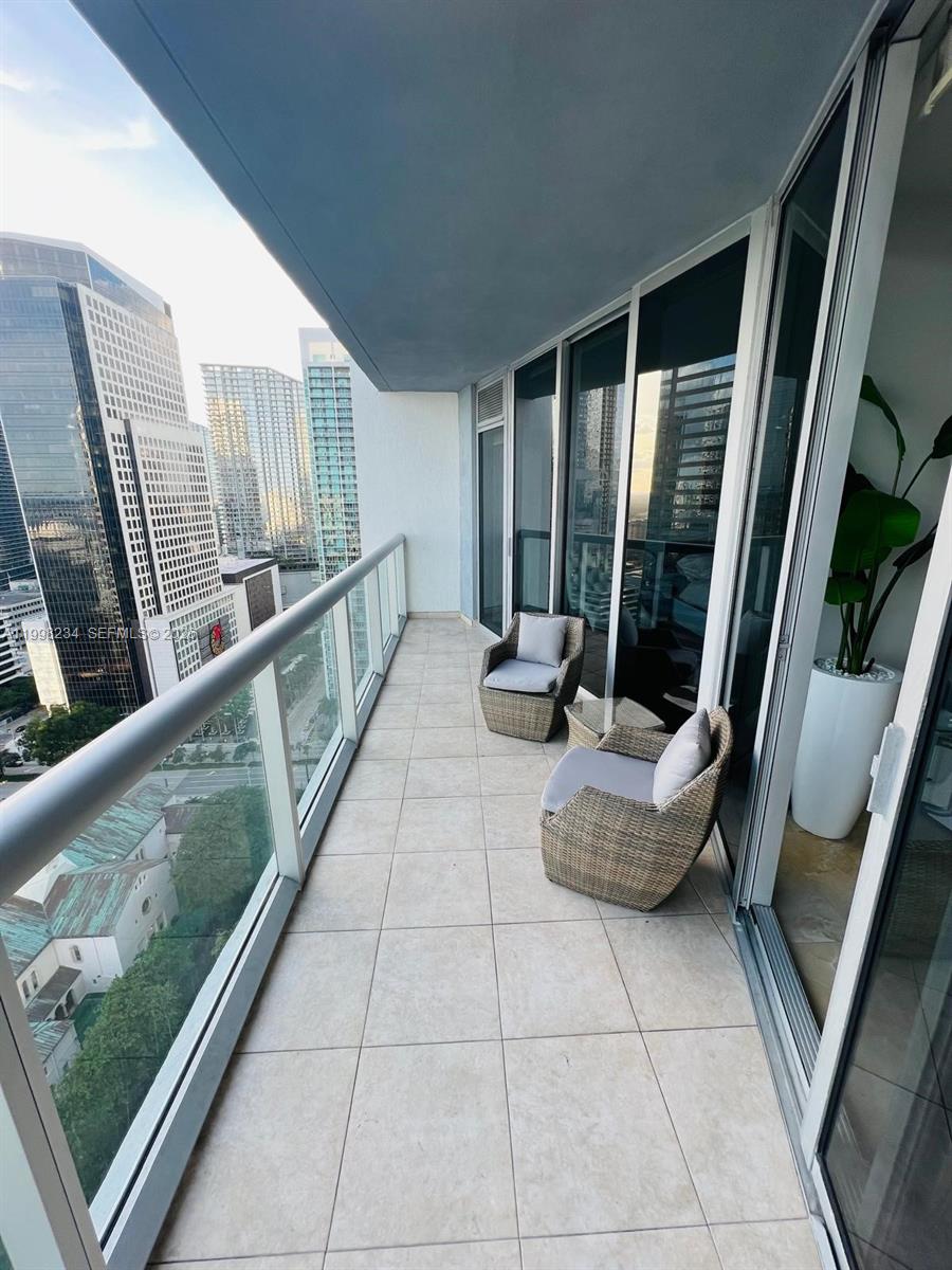 A high-rise balcony features tiled flooring, two wicker chairs, and a glass railing overlooking city buildings — 495 Brickell ave # 2707, Miami FL 33131, MIAMI, FL