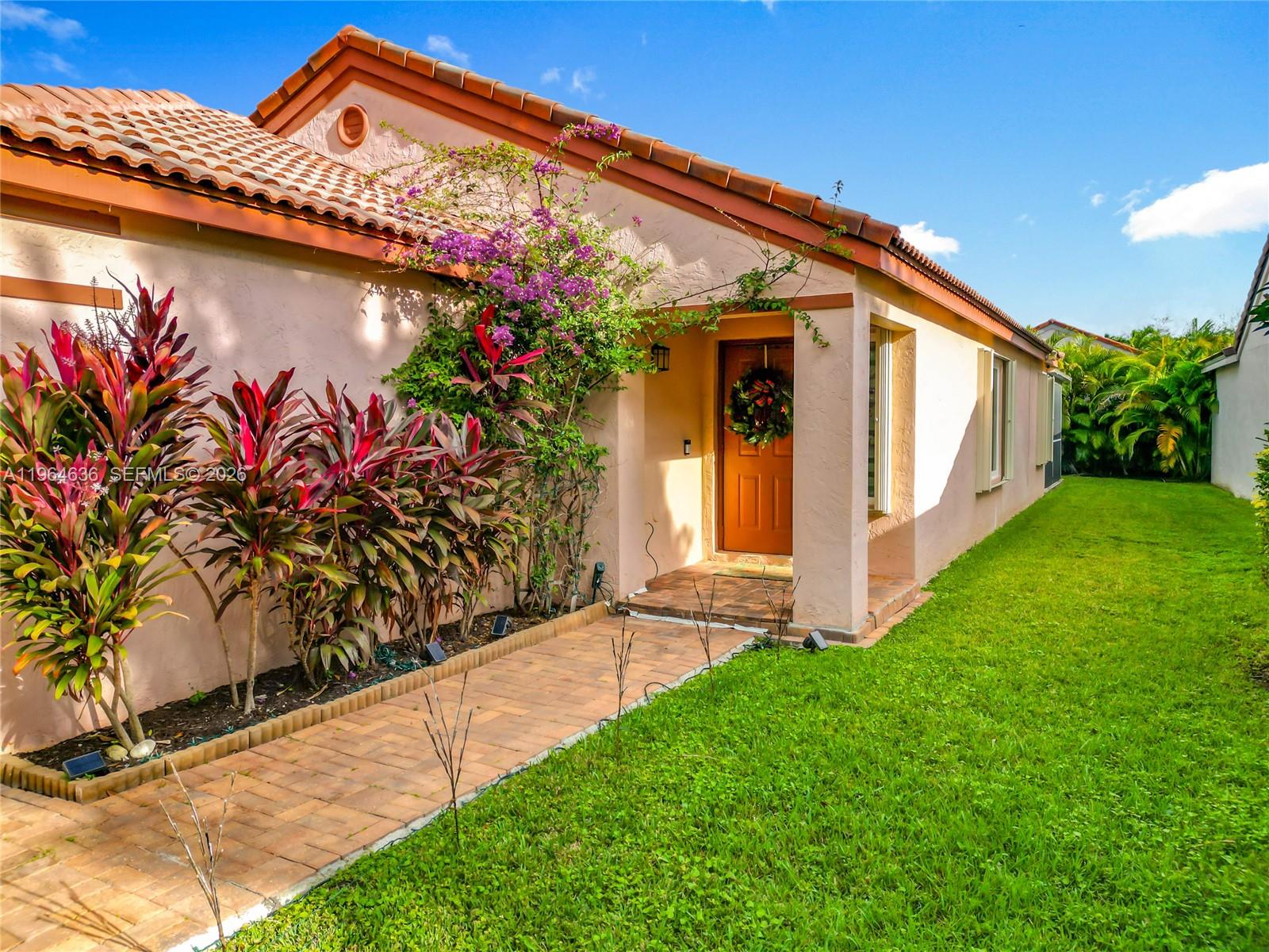 A peach-colored house features a brick walkway, a brown front door, and vibrant landscaping — 1536 Presidio Dr, Weston FL 33327, WESTON, FL
