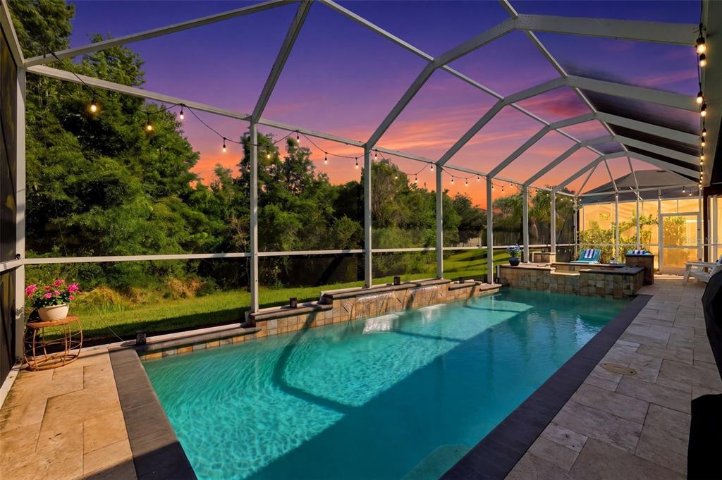 A screened-in swimming pool with a stone water feature and patio at sunset — 4107 WINDCREST DRIVE, WESLEY CHAPEL, FL