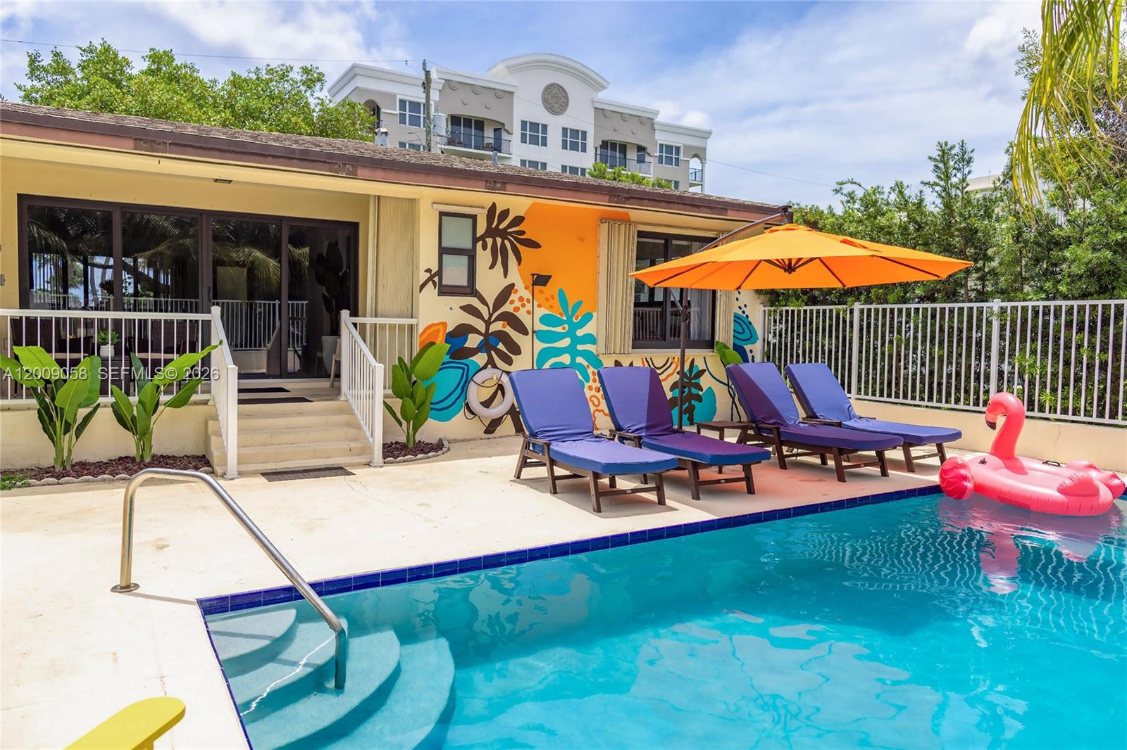A backyard swimming pool features four lounge chairs, an orange umbrella, and a mural wall — 155 SE 19th Ave # 0, Deerfield Beach FL 33441, DEERFIELD BEACH, FL
