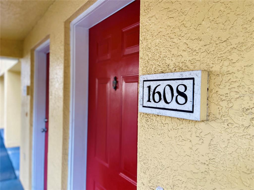 A red door with a white address plaque reading 1608 on a yellow stucco wall — 1608 LEGACY PARK DRIVE UNIT 1608, MAITLAND, FL