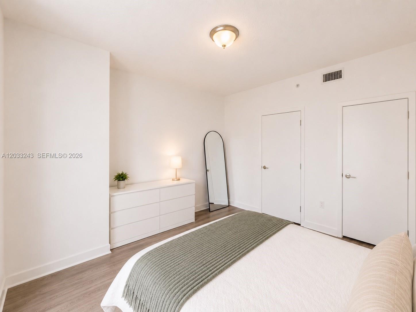 A bedroom features white walls, light wood flooring, a white dresser, and two closed doors — MIAMI, FL