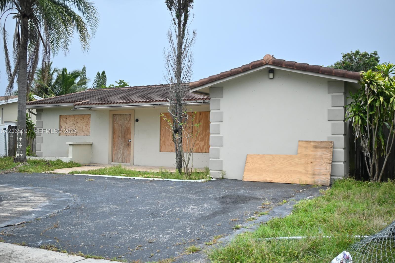 A single-story house with boarded-up windows and a door, featuring a tiled roof and driveway — HOLLYWOOD, FL