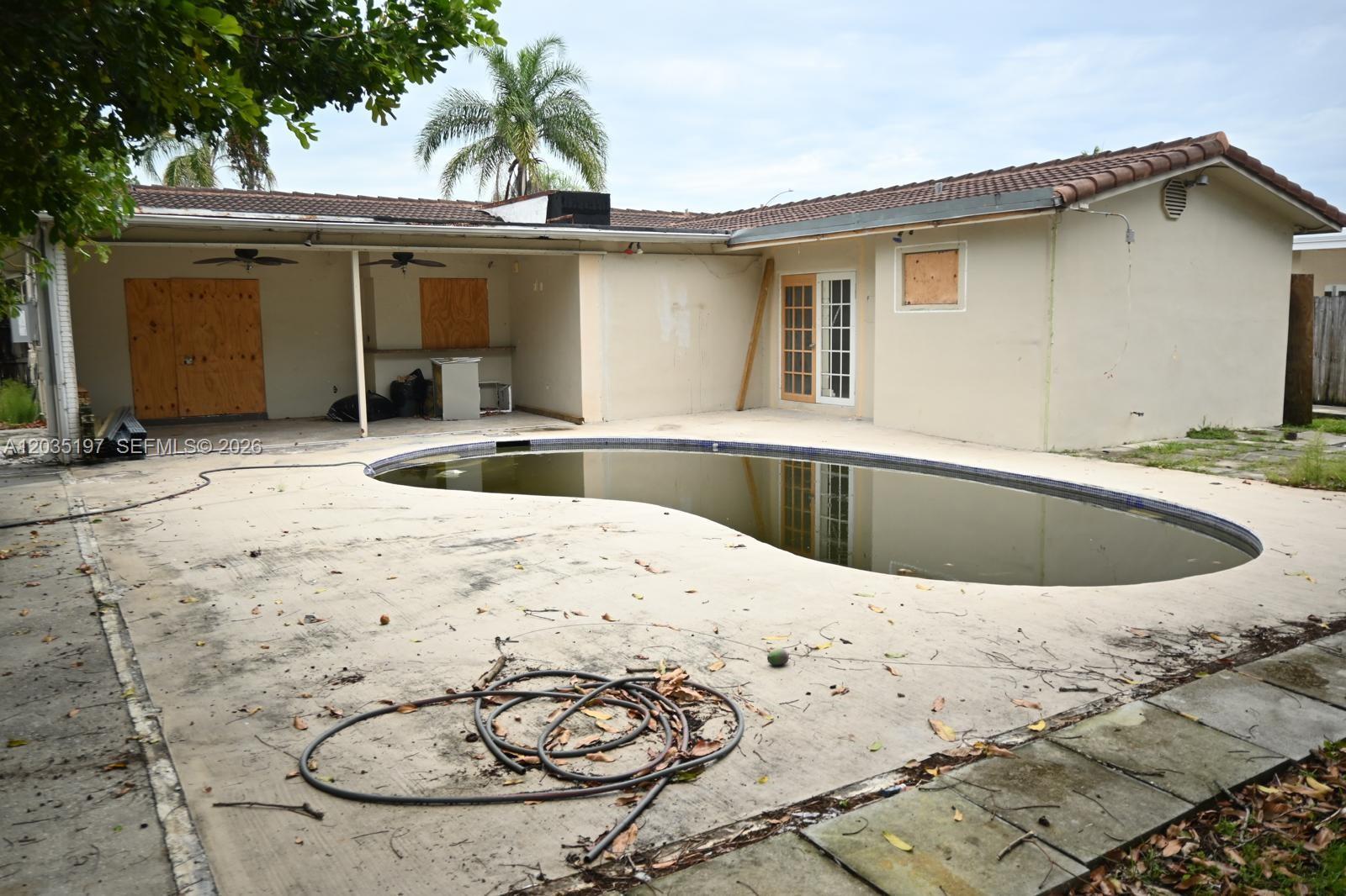 A backyard features a kidney-shaped pool, a covered patio, and a house with boarded windows — HOLLYWOOD, FL