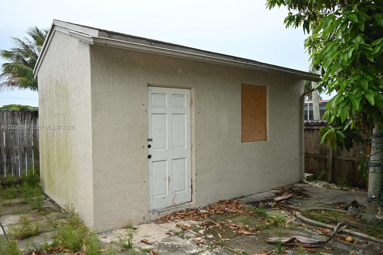 A detached, beige stucco shed with a white door and a boarded-up window — HOLLYWOOD, FL