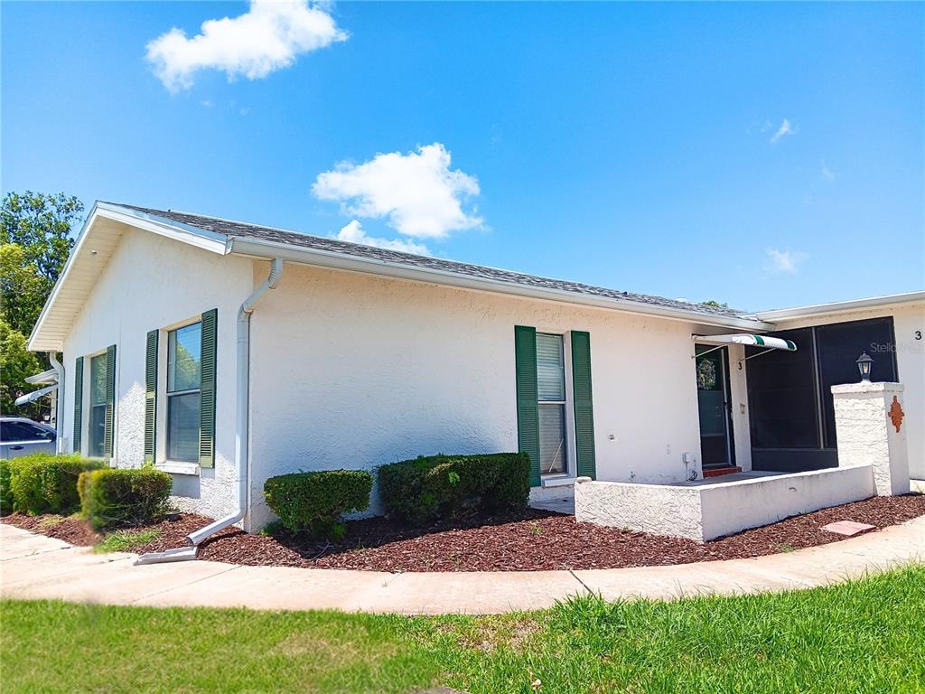 A single-story white stucco house features green shutters, a small front patio, and landscaping — 8150 BRADDOCK CIRCLE UNIT 3, PORT RICHEY, FL