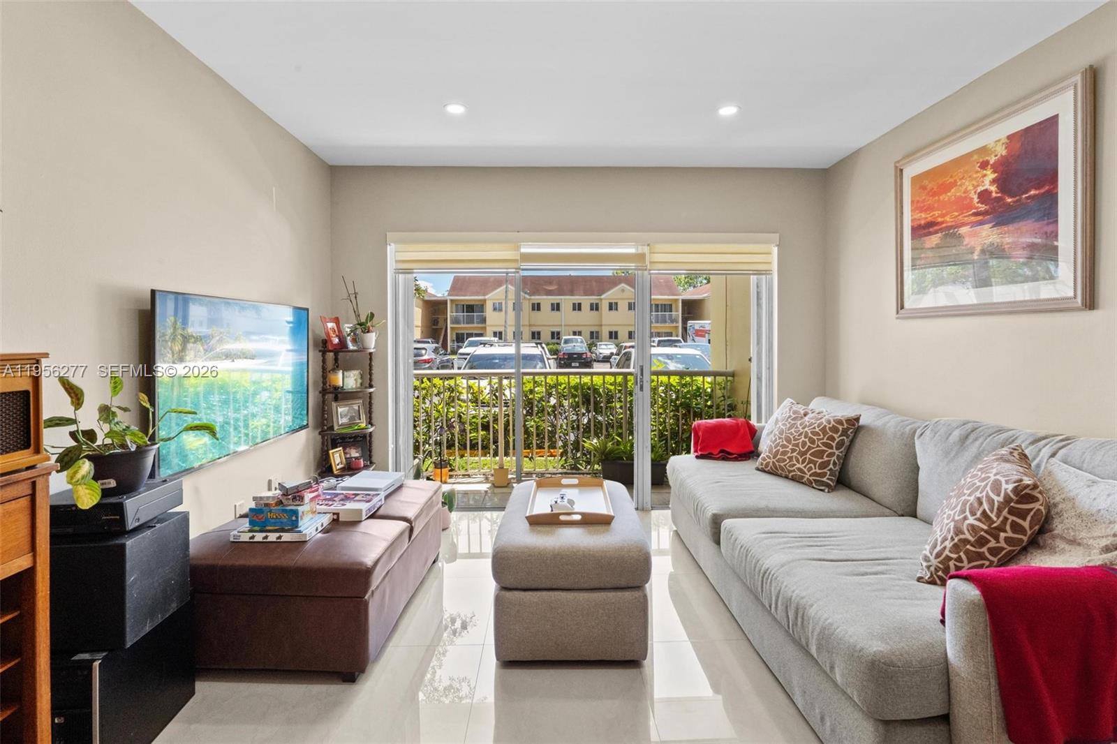 A living room features a gray sofa, a brown ottoman, a television, and a balcony view — 13810 SW 112th St # 102, Miami FL 33186, MIAMI, FL