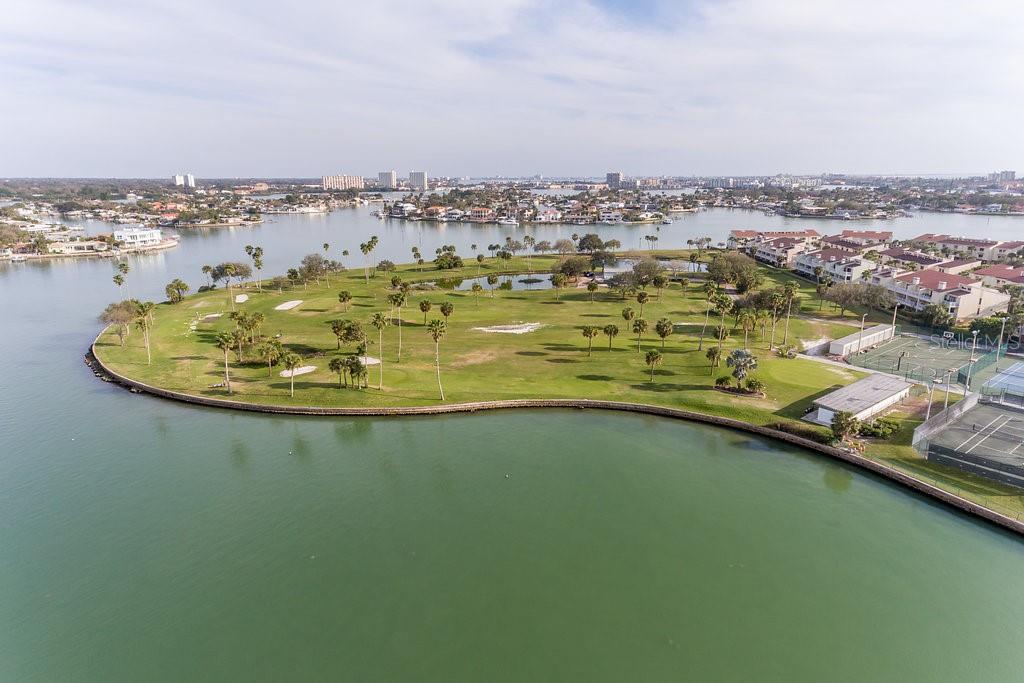 An aerial view of a waterfront golf course with palm trees and nearby residential buildings — 10355 PARADISE BOULEVARD UNIT 103, TREASURE ISLAND, FL