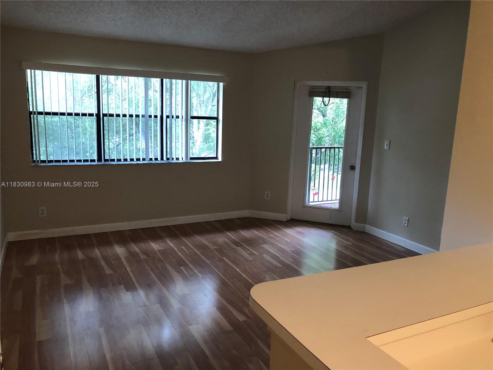 An empty living room features wood-look flooring, a large window with blinds, and a balcony door — 620 S Park Rd # 2, Hollywood FL 33021, HOLLYWOOD, FL