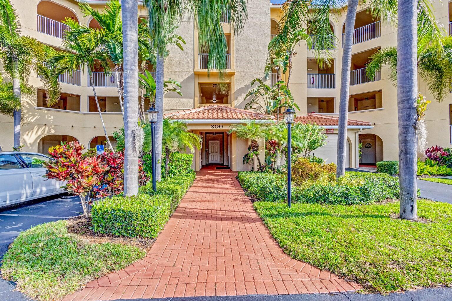A brick walkway leads to the entrance of a multi-story apartment building with palm trees — 300 Uno Lago Drive # 305, Juno Beach FL 33408, JUNO BEACH, FL