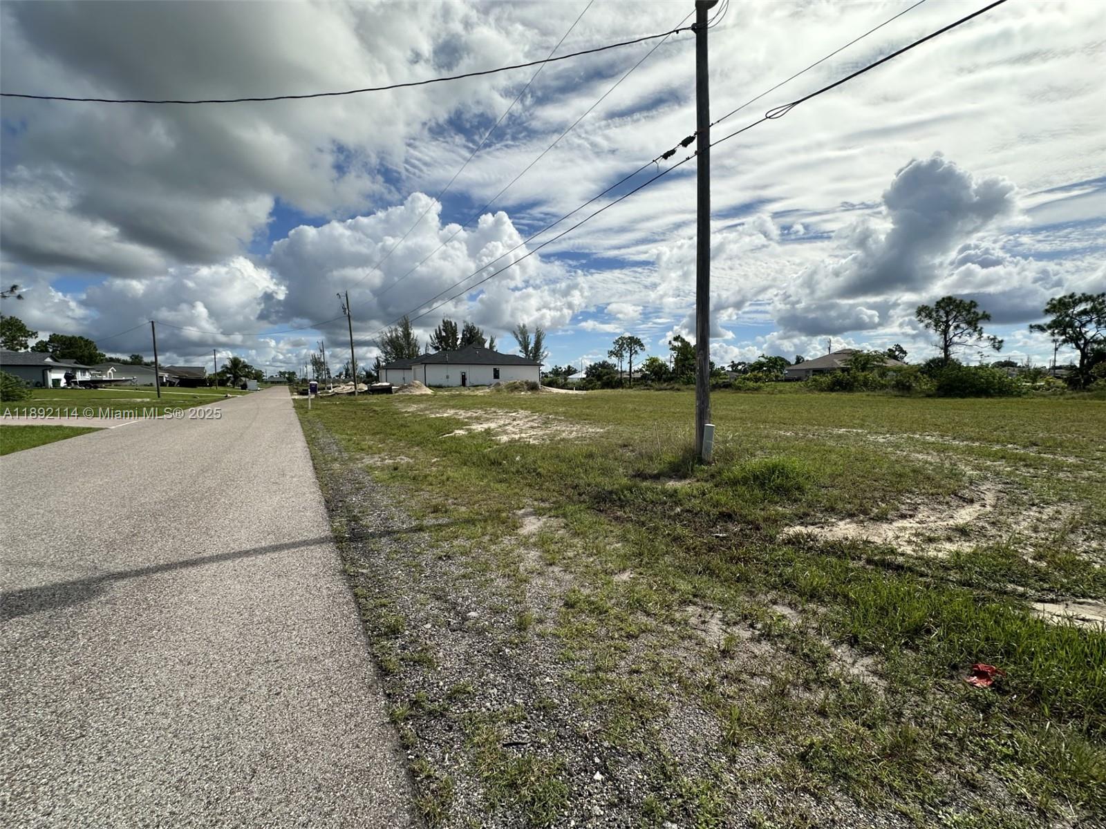 A paved road runs alongside a large, undeveloped grassy lot under a cloudy sky — 3527 NE 14 PL, Cape Coral FL 33993, CAPE CORAL, FL